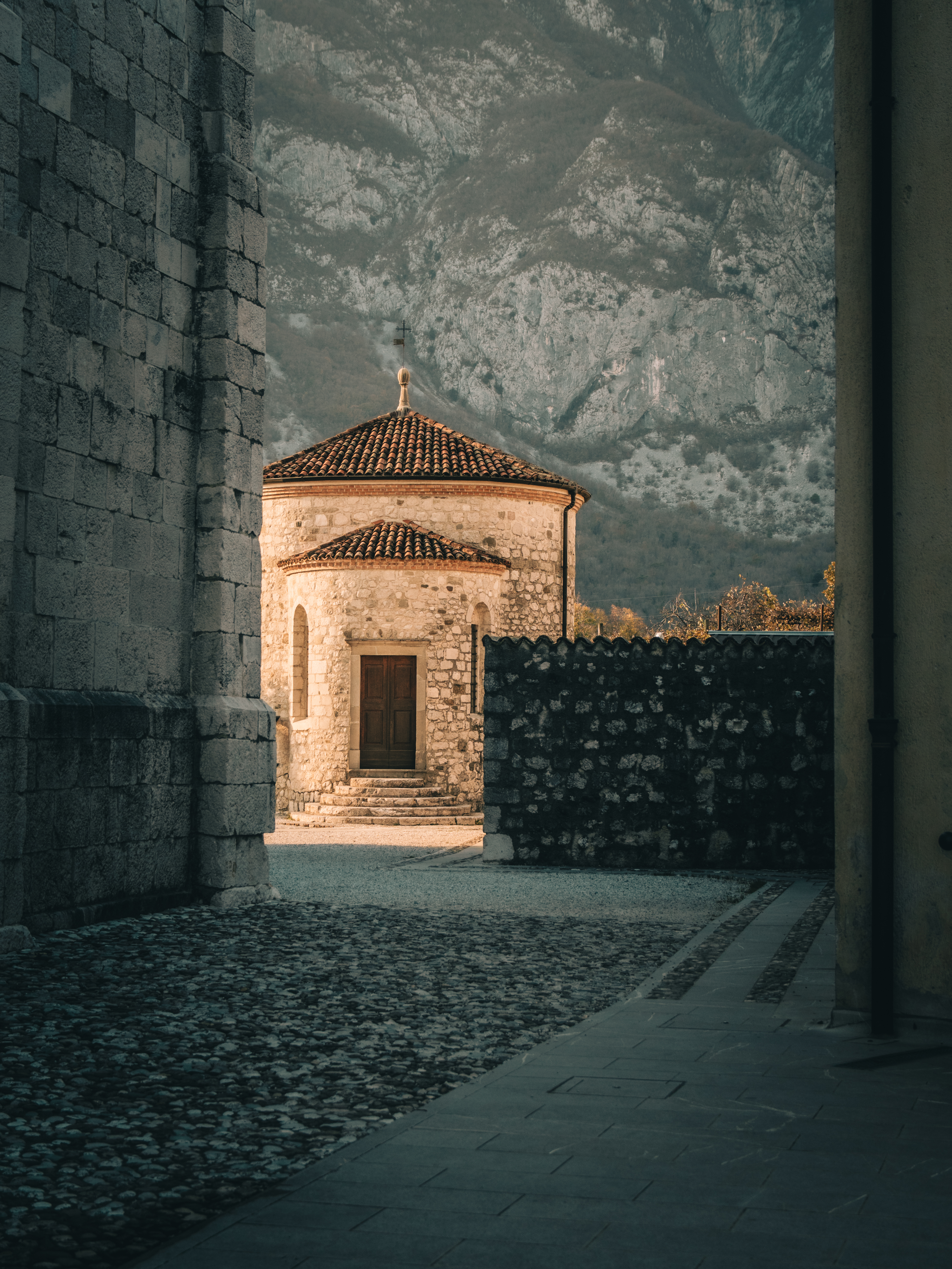 Kleine steinerne Kapelle mit rundem Dach und Kreuz, eingerahmt von zwei dunklen Mauern. Der Innenhof liegt im Schatten, die Kapelle im warmen Licht, dahinter rauge Berge. In Venzone, Italien.